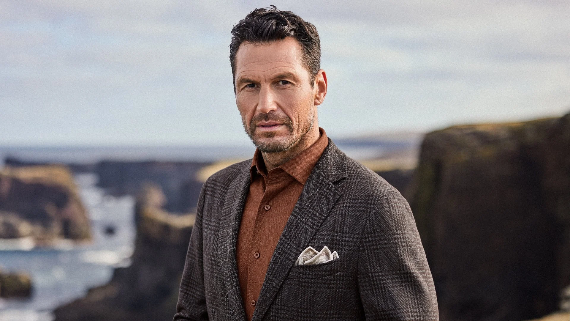 A man in a brown jacket and open collar standing on a grassy cliff edge above the sea