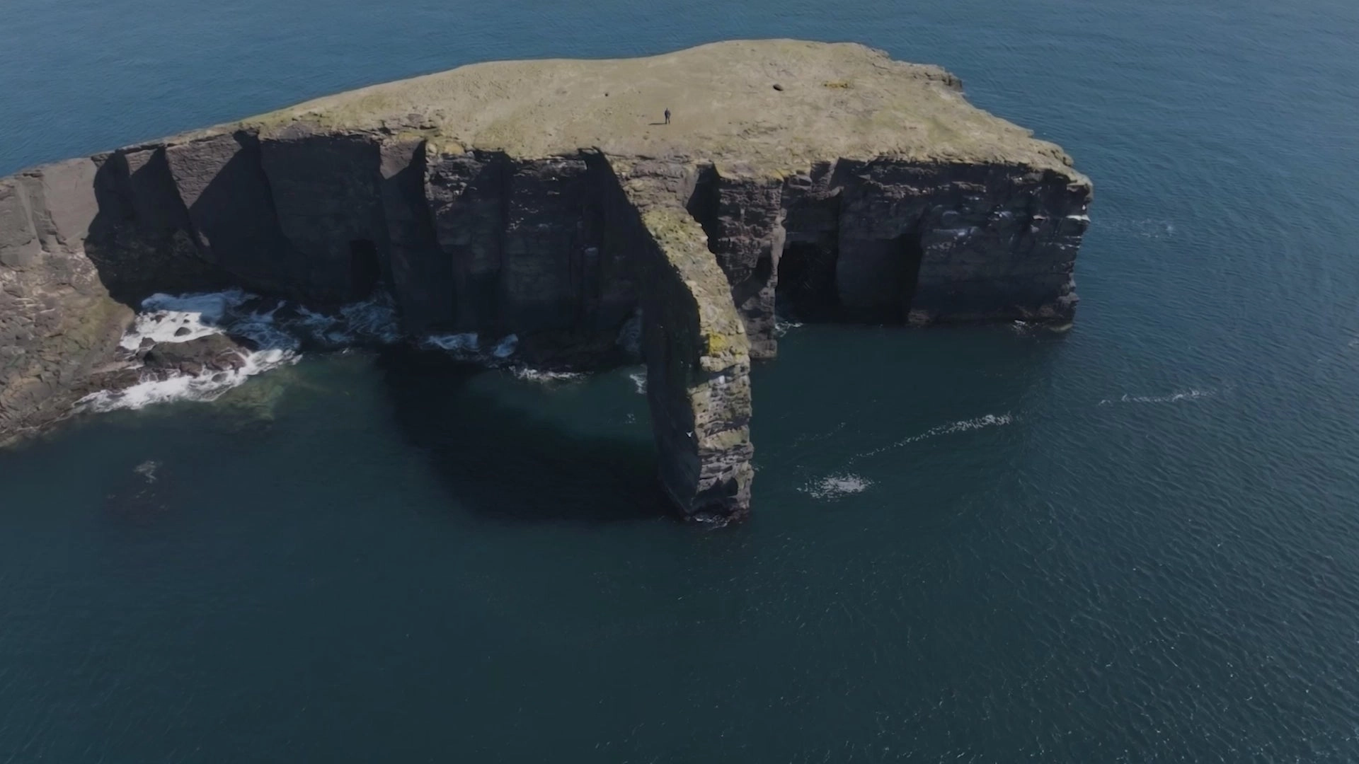 Aerial view of a dark rock stack off a coastline in blue-green water