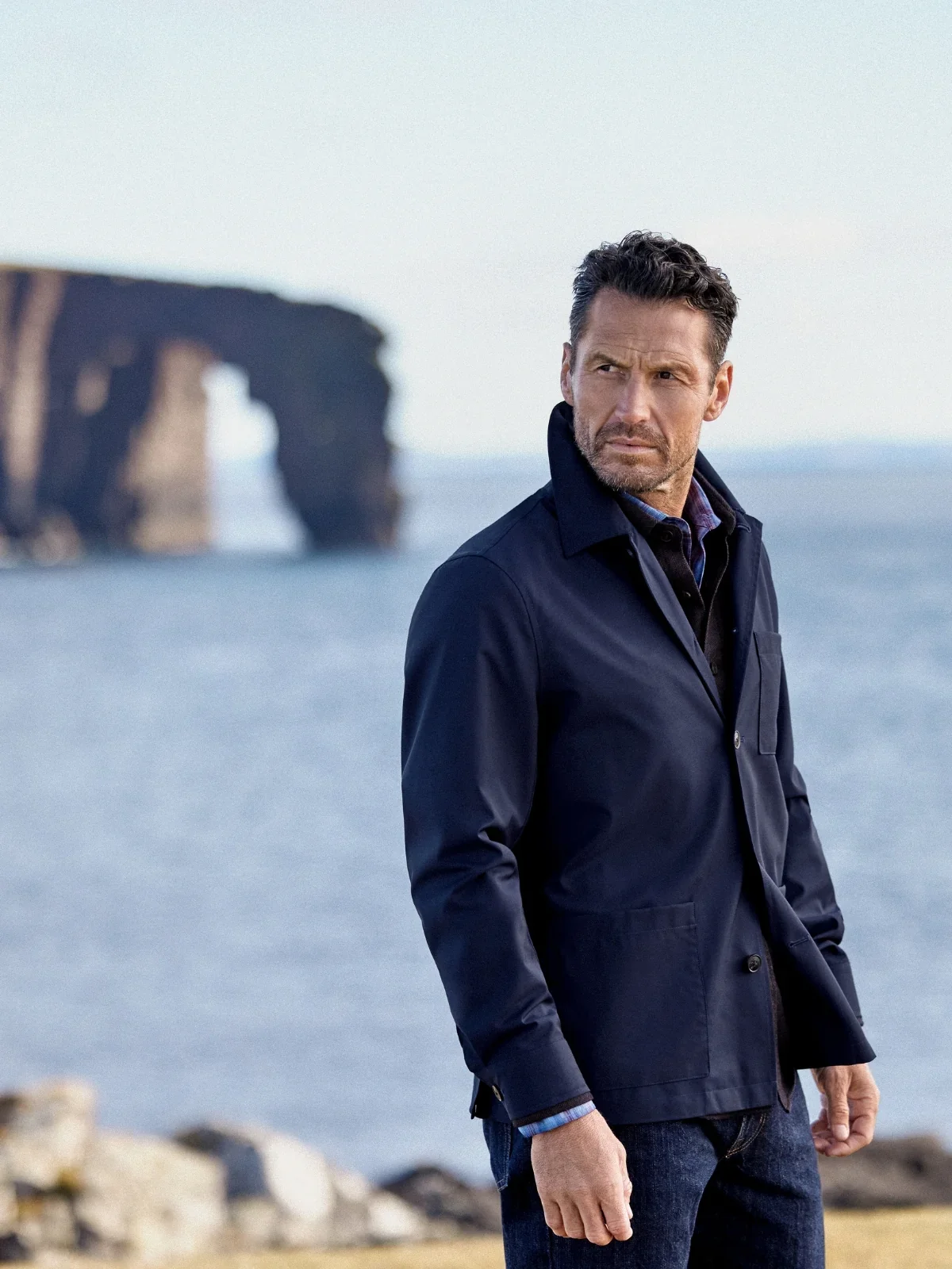 A man in a navy chore jacket in front of a sea arch on Shetland’s coast