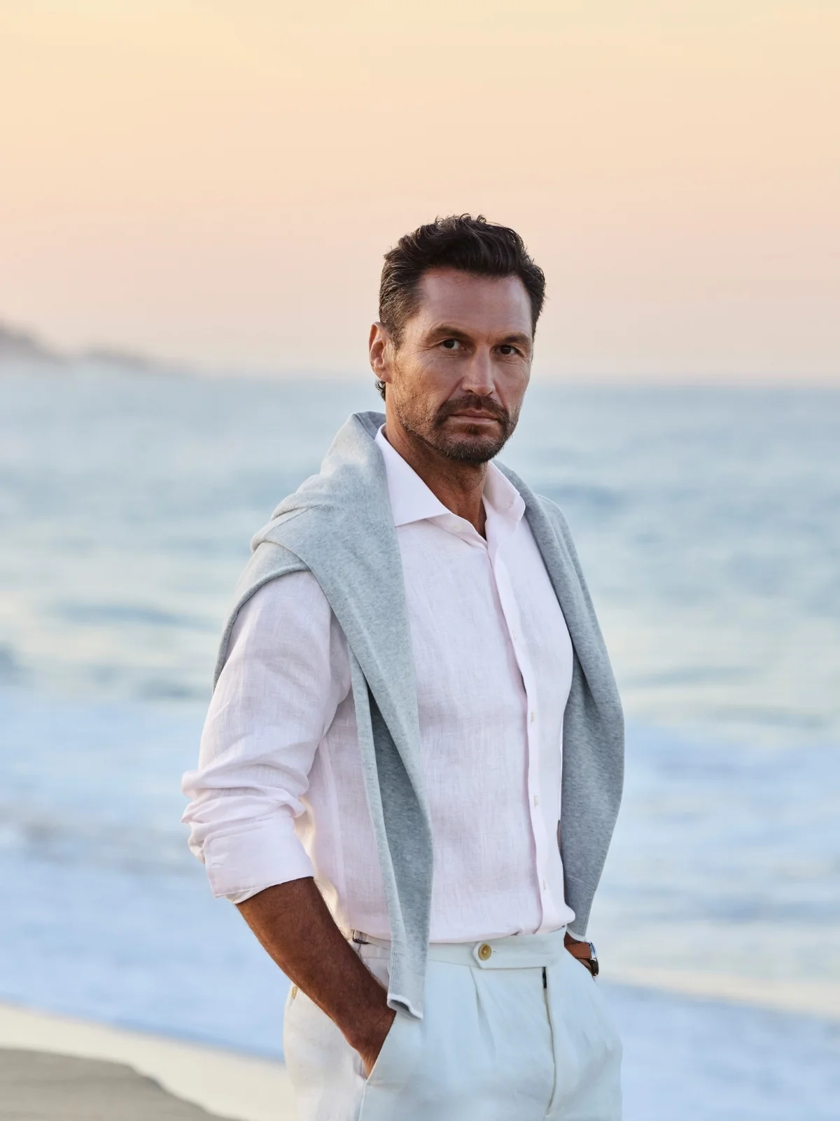A man in a white open-collar shirt with a grey sweater over his shoulders on a beach at dusk